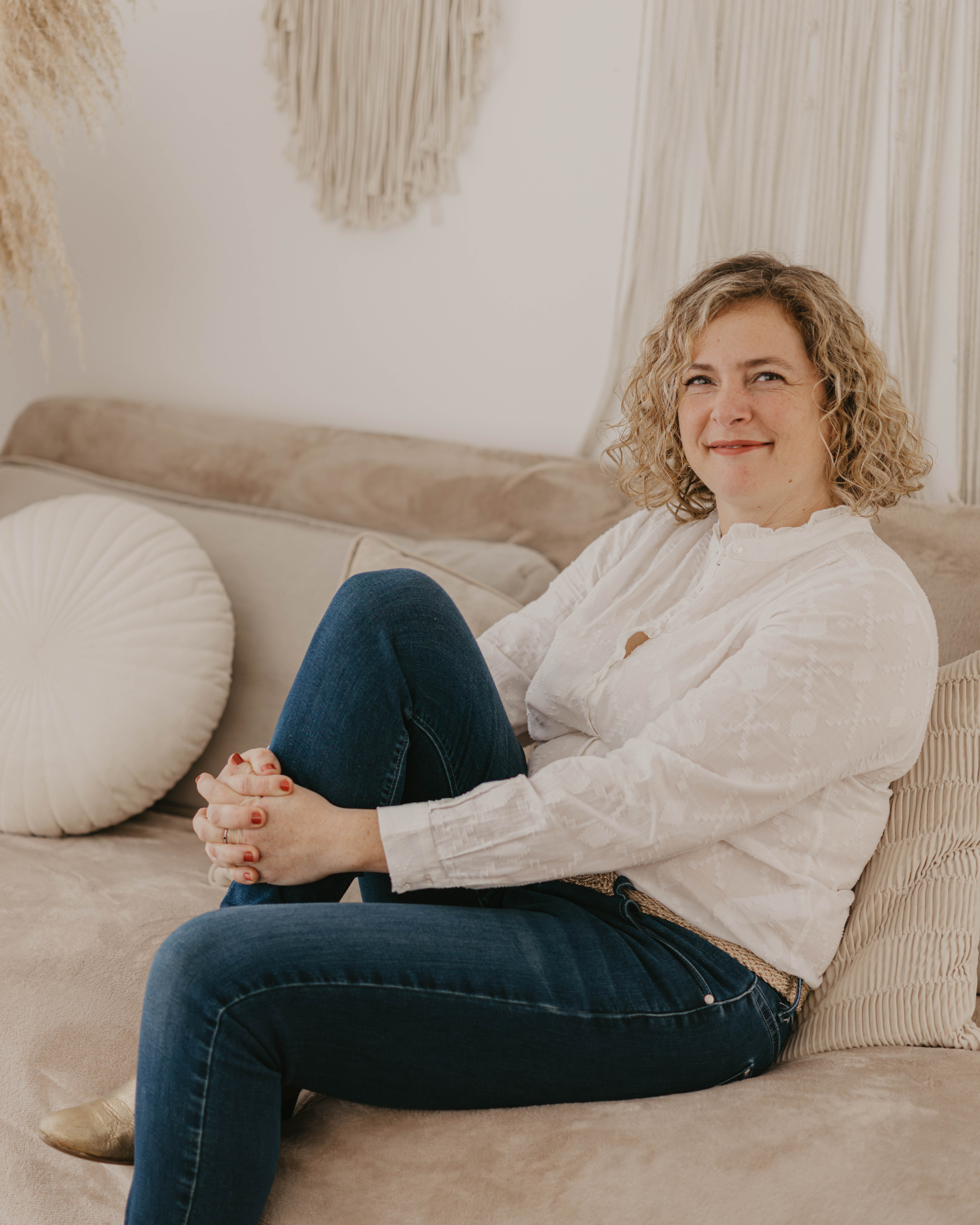A person with curly hair sits on a beige couch, wearing a white shirt and jeans, with a calm expression and decorative pillows around.